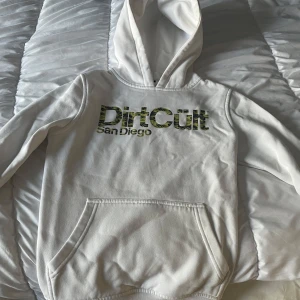 Vit hoodie från DirtCult San Diego - Säljer en vit hoodie från DirtCult San Diego med kamouflagemönstrad logga på bröstet. Tröjan har huva och en stor magficka framtill. Perfekt för dig som gillar streetwear och vill ha en bekväm och stilren look.