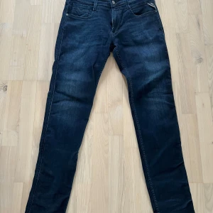 Mörkblå Replay Anbass jeans - Säljer ett par mörkblå Replay Anbass jeans med klassisk femficksdesign och snygga slitningar. Jeansen har normal passform och raka ben, med Replay-logga på bakfickan och läderpatch i midjan.