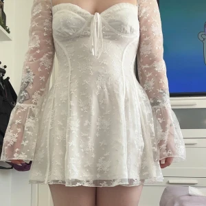 Vit blommig miniklänning från Boohoo - Superfin vit miniklänning från Boohoo med transparenta långa ärmar och blommigt mönster. Klänningen har en söt knytdetalj framtill och är figurnära upptill med lätt utsvängd kjol. Perfekt för sommarens tillfällen! Säljer åt en kompis som minst vill ha 300kr