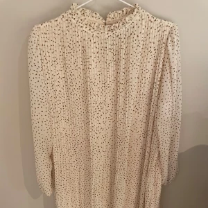 Beige prickig långklänning med volang - Superfin beige långklänning med små bruna prickar och plisserad struktur. Hög hals med volangdetalj och långa ärmar. Perfekt för dig som gillar en romantisk och trendig stil. Kommer med ett skärp