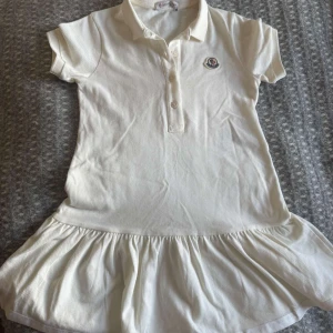 Vit kort klänning från Moncler för barn - Supersöt vit kort klänning från Moncler med pikékrage, knappar framtill och volang nertill. Klänningen har korta ärmar och broderad Moncler-logga på bröstet. För barn 6år aldrig använd 