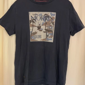 Mörkblå t-shirt från Morris - Snygg mörkblå t-shirt från Morris med tryck framtill som visar palmer, båtar och texten 'Bleu Navy'. Klassisk passform och rund halsringning. Perfekt för dig som gillar stilrena och coola tryck. Gjord i mjuk bomull för skön känsla.