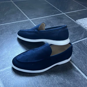 Mörkblå loafers - Snygga mörkblå loafers i mocka med stilren design och vit platt sula. Skorna har en klassisk rund tå och är perfekta för dig som gillar en clean och modern look. Skick 10/10, dem är helt nya.