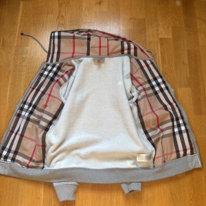 Grå hoodiejacka från Burberry M - Snygg grå hoodiejacka från Burberry med klassiskt rutigt foder i beige, svart och rött. Jackan har dragkedja, huva med snörning och två stora fickor framtill. Utsidan är i mjuk bomull och insidan har mysigt teddyfoder. Perfekt för dig som gillar streetstyle med lyxig touch. Liten i storlek, pris kan diskuteras
