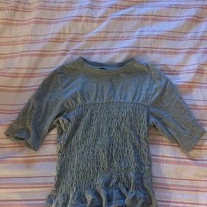 Grå smockad topp från H&M Divided XS - Grå topp från H&M Divided i storlek XS. Toppen har smockad midja och volangkant nertill, med korta ärmar och rund halsringning. Mjuk bomullskänsla och enkel, stilren look som passar till jeans eller kjol.