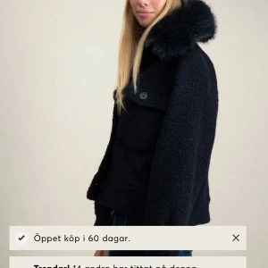 Svart teddyjacka med fuskpäls-krage - Snygg svart teddyjacka med stor fluffig fuskpäls-krage. Jackan har knäppning framtill med stora knappar och två bröstfickor. Modellen är boxig och lite croppad, perfekt till jeans och mössa. Materialet är mjukt och ger en cool vibe. jackan är från fina tricot