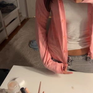 Ljusrosa kofta med knappar - Säljer en söt ljusrosa kofta med knappar framtill. Koftan har långärmade ärmar och en klassisk passform som är enkel att styla över ett linne eller t-shirt. Perfekt för lager-på-lager och ger en fräsch vibe till din outfit.