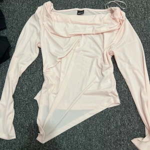 Ljusrosa långärmad topp Gina Tricot - Säljer en ljusrosa långärmad topp från Gina Tricot med draperad halsringning och asymmetrisk nederkant. Toppen är i ett mjukt, stretchigt syntetmaterial och har en snygg, lite lös passform. Perfekt för dig som gillar trendiga och unika detaljer.