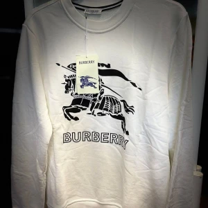 Vit Burberry sweatshirt med tryck - Snygg vit sweatshirt från Burberry med stort svart tryck av riddare och logga framtill. Tröjan har rund hals, långa ärmar och är gjord i mjuk bomull. Perfekt för dig som gillar klassisk stil med en modern twist.