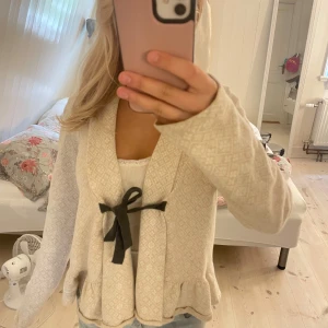 Odd Molly beige kofta med rosett - Superfin beige kofta från Odd Molly med diskret mönster, broderad logga på ryggen och svart rosett framtill. Koftan har lång ärm, volangkant nertill och är i mjukt stickat material. Perfekt för lager-på-lager och chill vibes.