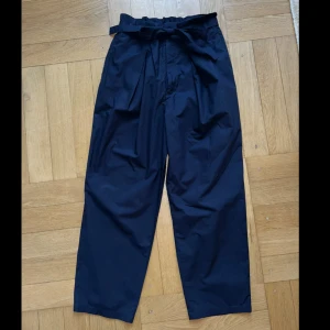 Mörkblå byxor med knyt från Slobe Iéna - Mörkblå byxor från Slobe Iéna med hög midja och knytband i midjan. Byxorna har raka, lite vidare ben och är tillverkade i en blandning av polyester, bomull, rayon och cupro. Snygg papperspåse-midja och två bakfickor för en chill och trendig look.