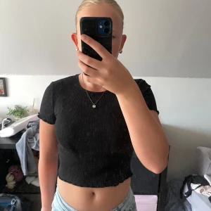 Svart ribbad croppad topp Gina Tricot - Säljer en svart croppad topp från Gina Tricot med ribbad struktur. T-shirten har korta ärmar och rund halsringning, perfekt för en chill och stilren look. Enkel att matcha med jeans eller kjol.