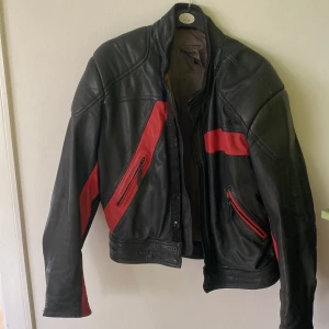Svart och röd mc jacka med dragkedjor - Cool svart mc skinnjacka med röda detaljer och dragkedjor på fickorna. Jackan har en sportig vibe med markerade axlar och röda inslag längs ärmar och bröst. Perfekt för dig som vill sticka ut med en edgy look. Passar dig som gillar streetstyle och klassiska bikerjackor.