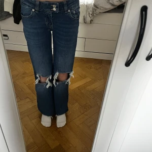 Blå bootcut jeans med slitna knän - Säljer ett par mörkblå jeans med bootcut passform och stora slitningar över båda knäna. Jeansen har klassisk femficksdesign, låg midja och är tillverkade i kraftig denim. Perfekta för en edgy och avslappnad stil. OBS! Dom är uppsydda några centimeter!