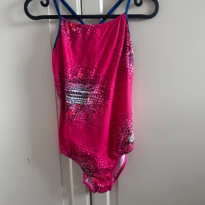 Rosa baddräkt från Arena med mönster - Snygg rosa baddräkt från Arena med coolt prickigt mönster i svart, vitt och färgglada detaljer. Smala blå axelband som korsas i ryggen ger en sportig vibe. Perfekt för simträning eller strandhäng.