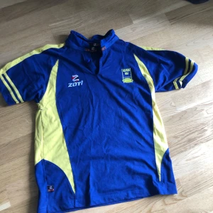 Blå och gul fotbollströja Zoti - Säljer en blå fotbollströja från Zoti med gula detaljer längs sidorna och ärmarna. Tröjan har korta ärmar, ståkrage och klubbmärke på bröstet. Perfekt för dig som gillar sportig stil och vill sticka ut på planen.