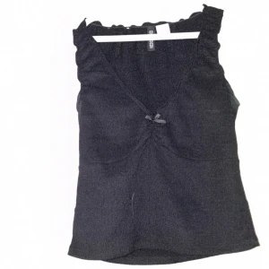 Svart linnetopp med rosett från H&M - Supersöt svart linnetopp från H&M med v-ringning och liten rosett framtill. Toppen har breda axelband och en lätt rynkad detalj vid bysten som ger en feminin touch. Perfekt att styla med jeans eller kjol för en trendig look.