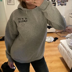 Grå hoodie från Gina Tricot med tryck - Grå hoodie från Gina Tricot med svart text 'Je m'en Amour' och en stjärna tryckt på både fram- och baksidan. Rätt så mjuk insida men är tvättad ett antal gånger, huva och klassisk passform. Köpt på Gina för ungefär 250kr om jag minns rätt! 