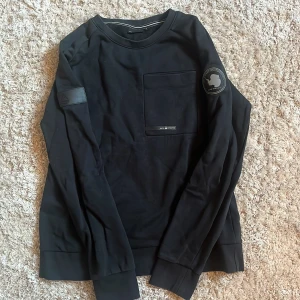 Svart långärmad tröja från Paul & Shark - Svart långärmad tröja från Paul & Shark med rund halsringning och stor bröstficka. Snygga patchar på ena ärmen och axeln ger en cool touch. Perfekt för dig som gillar stilrena och sportiga plagg.