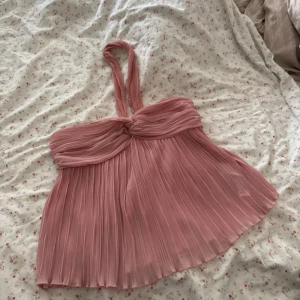 Plisserad rosa halterneck linnetopp - Supersöt rosa linnetopp med plisserat tyg och halterneckband. Toppen har twistad detalj framtill och ett flowy, luftigt fall. Perfekt för varma dagar när du vill ha något lätt och snyggt. Materialet känns mjukt och svalt mot huden.