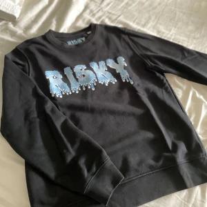 Svart sweatshirt från Risky med tryck - Svart sweatshirt från Risky med stort blått och vitt drip-tryck över bröstet. Tröjan har rund halsringning, långa ärmar och ribbade muddar. Perfekt för dig som vill ha en skön cool hoodie nu till hösten som passar till det mesta 