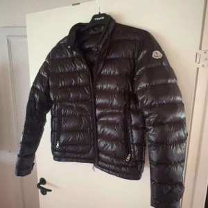 Svart Moncler dunjacka med dragkedja - Snygg svart dunjacka från Moncler med glansig finish och klassisk quiltad design. Jackan har hög krage, två fickor med dragkedja och Moncler-logga på ärmen. Perfekt för kalla dagar när du vill hålla stilen.