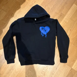 Svart hoodie med blått tryck - Svart hoodie med huva och känguruficka. På ryggen står det 'LOVE' i blått med stadssilhuett och samma hjärta. Perfekt för dig som gillar streetwear och unika prints.