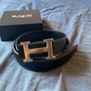 Svart skärp med H-spänne i skinn - Stilrent svart bälte i skinn med vit kontrastsöm och ett stort, silvrigt H-format metallspänne. Perfekt accessoar för att lyfta din outfit med en lyxig touch. Levereras i original ask med logga.