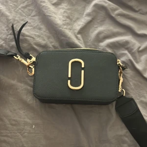 Svart axelväska från Marc Jacobs - Snygg svart axelväska från Marc Jacobs med guldiga detaljer och bred justerbar axelrem. Väskan har två dragkedjor upptill, stilren rektangulär form och ikonisk logga framtill. Tillverkad i skinn med struktur och har en praktisk ficka på baksidan.