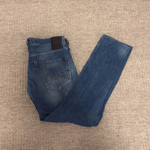 Blå Replay jeans - Säljer ett par klassiska blå jeans från Replay med snygga slitningar och raka ben. Jeansen har fem fickor, normal passform och detaljerade sömmar. Perfekta för dig som gillar en tidlös och avslappnad stil.