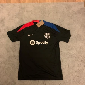 FC Barcelona svart Nike fotbollströja - Säljer en svart FC Barcelona fotbollströja från Nike med klubbmärke och Spotify-logga på bröstet. Tröjan har korta ärmar och detaljer i rött och blått på axlarna. Materialet är lätt och andas, perfekt för träning eller match. Matchande shorts medföljer, även de med klubbmärke.