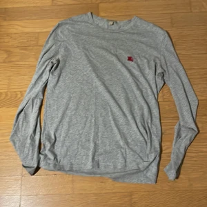 Grå långärmad tröja från Burberry - Snygg grå långärmad tröja från Burberry i 100% bomull. Tröjan har en klassisk passform och är dekorerad med Burberrys ikoniska röda broderade ryttare på bröstet. Perfekt för dig som gillar stilrena och tidlösa plagg.
