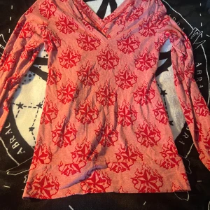 Rosa långärmad tröja med rött mönster - Långärmad rosa tröja med unikt rött mönster av stjärnor och eldflammor. Tröjan har v-ringad hals och är gjord i mjukt bomullsmaterial. Perfekt för dig som vill sticka ut med coola detaljer och bekväm passform.