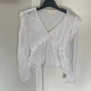 Vit volangblus från Gina Tricot - Supersöt vit blus från Gina Tricot med v-ringning och volangdetaljer framtill och på axlarna. Långärmad och i lätt, luftig bomull som ger en romantisk vibe. Perfekt att styla med jeans eller kjol för en fräsch look.