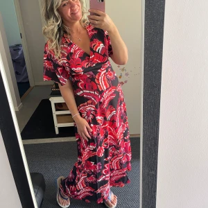 Rödblommig långklänning med v-ringning - Säljer en maxiklänning med röd- och vitblommigt mönster på svart botten. Klänningen har v-ringning, korta vida ärmar och markerad midja. Tyget är lätt och flowy, perfekt för varma dagar. Supersnygg och färgstark look!Helt ny med etikett 