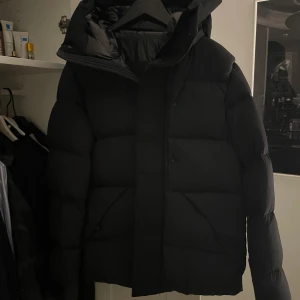 Moncler vinterjacka - Säljer en svart pufferjacka med stor huva och breda, quiltade partier. Jackan har dragkedja och knappar framtill samt två snedställda fickor. Perfekt för kalla dagar och riktigt skön att bära.