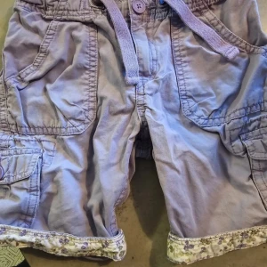 Ljuslila shorts med blommig kant L.O.G.G - Säljer ett par ljuslila shorts med flera fickor, snörning i midjan och blommigt tyg på insidan av linningen och bensluten. Shortsen har coola detaljer som knappar och praktiska fickor, perfekt för dig som gillar en avslappnad stil med en söt twist.