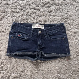 Mörkblå jeansshorts från Hollister - Snygga mörkblå jeansshorts från Hollister med råa kanter och klassisk femficksdesign. Shortsen har låg midja och en liten Hollister-logga på fickan fram. Perfekta för varma dagar och har en avslappnad, trendig vibe. De är i strl W 23 är i superbra skick men har en reva på märket som man ser på bild 3, det är dock inget man tänker på🥰