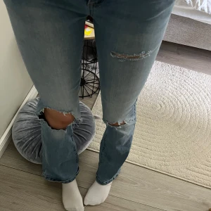 Ljusblå bootcut jeans med slitningar - Säljer ett par ljusblå jeans med bootcut-modell och snygga slitningar vid knäna. Jeansen har klassisk femficksdesign, normal passform och är tillverkade i mjukt bomullstyg. Perfekta för dig som gillar en avslappnad och trendig look.