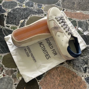 Golden Goose Superstar skor Storlek 42 - Golden Goose Superstar sneakers i vitt skinn med grå mockadetaljer och stjärna på sidan. Grå snörning, mörkblå häl och beige sula med vintagekänsla. Klassisk låg modell med platt sula och rund tå. Snyggt slitna detaljer för en cool look. Kommer utan skokartong