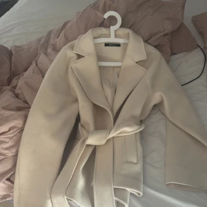 Beige kappa  - Stilren beige kappa från Gina med klassisk krage och bälte i midjan. är perfekt för dig som vill ha en clean och trendig look. Tidlös design som funkar till många outfits.