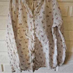 Blommig vit blus från Newbie - Supersöt vit blus från Newbie med små blommor i rosa och grönt. Blusen har långa ärmar, knappar framtill och volangdetalj vid halsen. Perfekt för dig som gillar romantisk och vintageinspirerad stil.