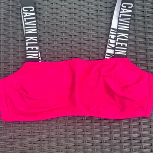 Rosa bikiniöverdel från Calvin Klein - Snygg rosa bikiniöverdel från Calvin Klein med breda axelband i svart och vitt med logga. Axelbanden är elastiska och ger en sportig vibe. Överdelen har en enkel, rak form och stängs med spänne bak. Perfekt för stranden eller poolen!