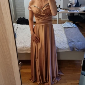 Champagnefärgad långklänning med strass - Elegant långklänning i champagnefärgad satin med offshoulder-skärning och vacker strassdekor längs urringningen. Klänningen har markerad midja, omlott-detaljer upptill och ett svepande, utsvängt kjolparti. Perfekt för bal eller festligheter med tjocka och ordentliga material. Köpt för lite över 4 000kr och använd en gång.