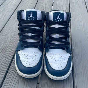Nike Air Jordan 1 Mid marinblå/vit (nypris 2400 sek) - Snygga Nike Air Jordan 1 Mid sneakers i marinblått och vitt skinn med svarta snören och klassisk Jumpman-logga på plösen. Perforerad tåbox och ikonisk Swoosh på sidan. Perfekta för dig som gillar streetstyle och vill sticka ut med en tidlös modell.