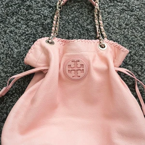  Tory Burch väska - Snygg ljusrosa axelväska från Tory Burch i mjukt skinn. Väskan har kedjehandtag med inslag av rosa läder, dekorativ snörning upptill och det klassiska Tory Burch-loggan framtill. Invändigt finns ett fack med dragkedja och logomönstrat foder.