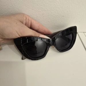 Svarta cat eye-solglasögon från Gufo - Snygga svarta solglasögon från Gufo i cat eye-modell med cool krokodilpräglad båge och trälook på skalmarna. Perfekta för dig som vill sticka ut med en unik accessoar och ge din outfit en edgy vibe.