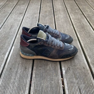 Valentino Garavani Rockrunner sneakers - Snygga Valentino Garavani Rockrunner sneakers i mörka toner med camouflage-mönster. Skorna har detaljer i mocka och skinn, grå snörning och en beige sula med nitar på hälen. Perfekta för dig som gillar streetstyle med lyxig touch.
