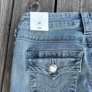 True Religion Joey Low Rise Flare Jeans - Snygga ljusblå jeans från True Religion, modell Joey med låg midja och utsvängda ben. Klassiska bakfickor med silverfärgade knappar, samt tydliga sömmar. Helt nya med prislapparna på!