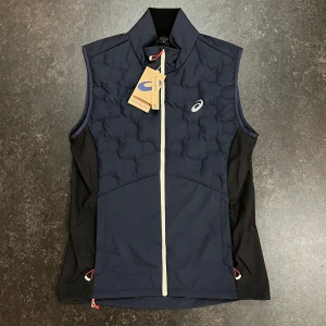 Mörkblå dunväst från Asics - Sportig mörkblå dunväst från Asics med quiltad framsida och vit dragkedja. Perfekt för löpning eller utomhusaktiviteter när det är kyligt.  Slim modell